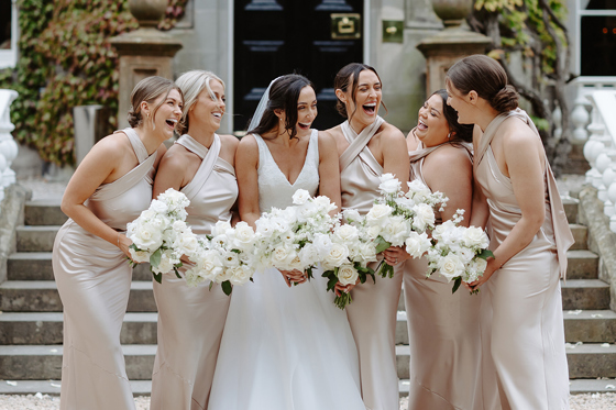 Bride and her five bridesmaids laugh together while holding white rose bouquets